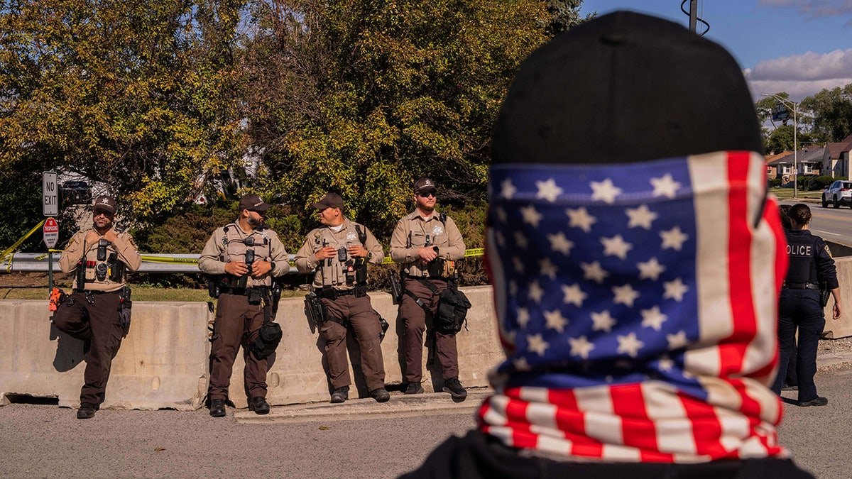 Un manifestant portant un couvre-visage avec un drapeau américain se tient en face des shérifs du comté de Cook devant un bureau de l'immigration et des douanes à Broadview, dans l'Illinois, le samedi 11 octobre 2025.