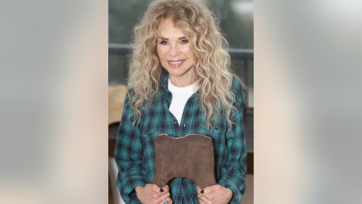 Dyan Cannon s'accroche à une chaise en bois et sourit dans un chemisier à carreaux vert et une chemise blanche.